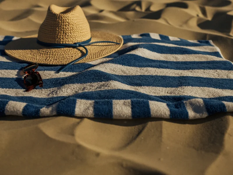 Gestreiftes Baumwoll-Strandtuch flach auf hellem Sand mit Sonnenbrille und Strohhut